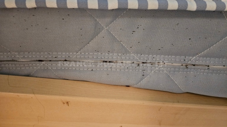 Flanc de matelas gris bleu matelassé, couture horizontale garnie de points noirs, cadre en bois clair dessous